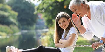 [GEN] An elderly woman leans in to whisper into a young girl's ear, seated on a park bench near a tranquil river, surrounded by lush greenery. No identifiable hearing aid brands or text present.