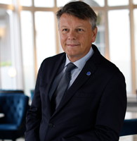 A man wearing a dark suit, tie, and a blue circular lapel pin is standing in a well-lit, modern lounge with blue chairs and large windows in the background.