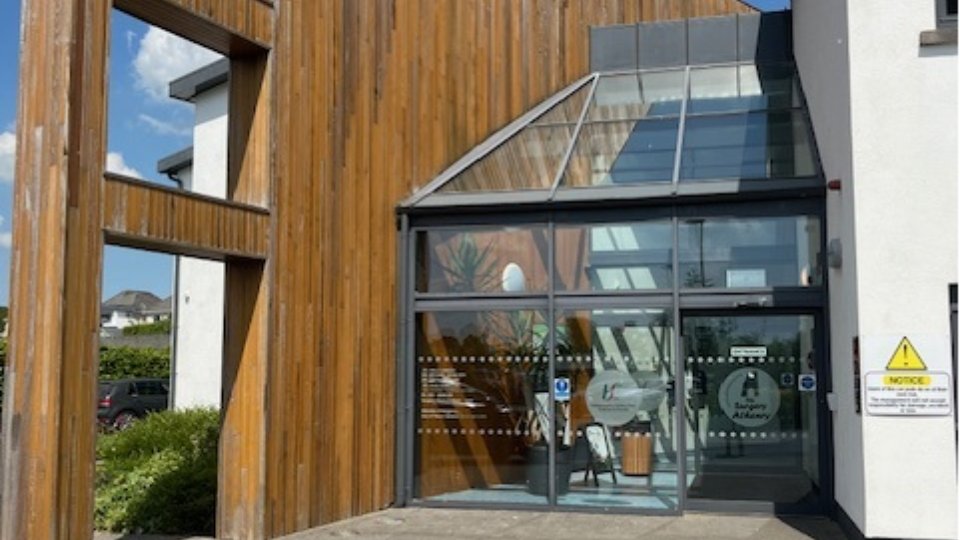 The main entrance of a modern building features wood paneling, glass doors, and a slanted glass roof. Text on the door includes 'Surgery At Albany.' A sign reads: 'NOTICE CCTV in operation.' The environment is sunny with shrubs and parked cars nearby.