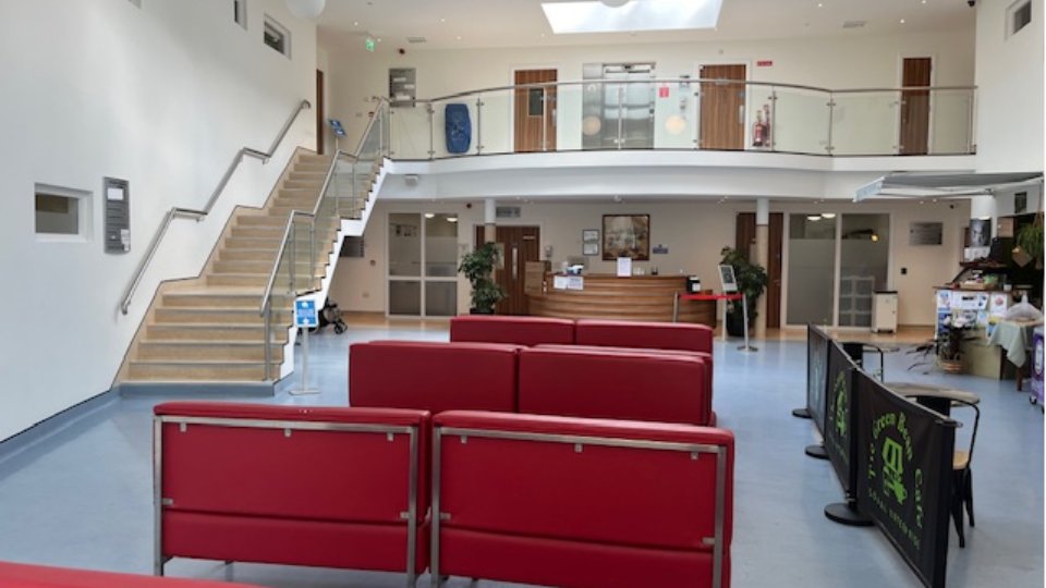 Red cushioned chairs face a wooden reception desk in a bright, spacious hearing clinic lobby with stairs, glass railings, and plants. Signs for 'The Green Bean Café' are visible. No text found indicates specific hearing aid brands.