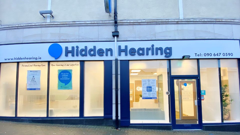 Hidden Hearing storefront features a white and blue sign reading 'Hidden Hearing,' with the website www.hiddenhearing.ie and phone number Tel: 090 647 0590. Posters promote hearing services like personalized care.