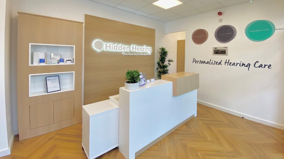 A modern hearing clinic reception area features a wooden desk with plants and brochures. The wall displays 'Hidden Hearing – Your hearing is our expertise' and 'Personalised Hearing Care' alongside decorative branding elements.