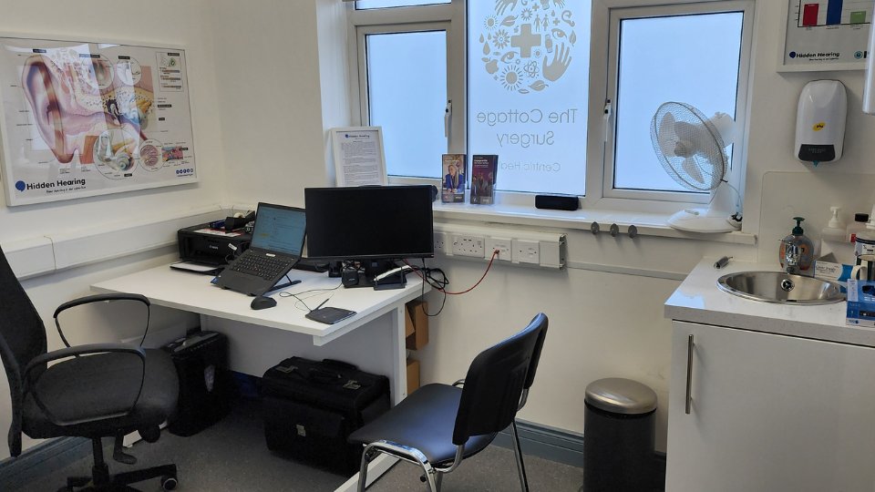 A hearing clinic room featuring a desk with a laptop, monitor, and audiology equipment, alongside chairs and a sink. A Hidden Hearing poster displaying an ear diagram hangs on the wall. Text on the window reads, 'The Cottage Surgery,' promoting its healthcare context.