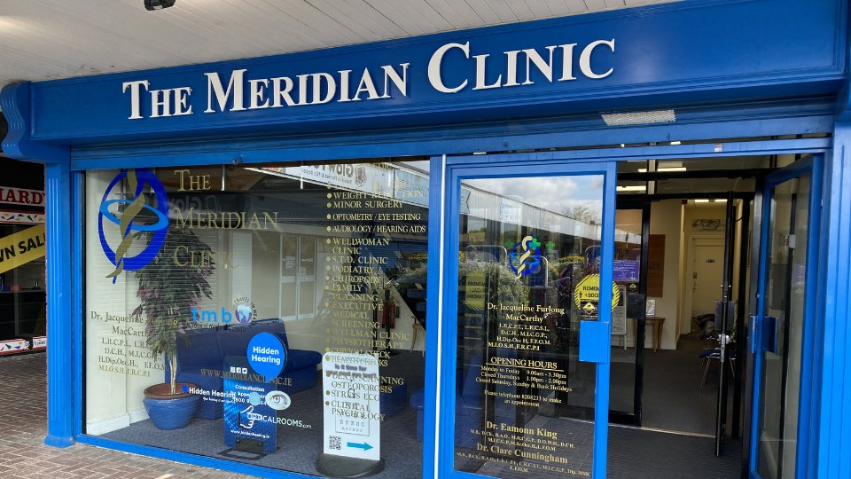 [GEN] The Meridian Clinic storefront features bold blue signage, glass doors, and windows displaying services like audiology, hearing aids, and eye testing. A Hidden Hearing kiosk promotes hearing care solutions inside.