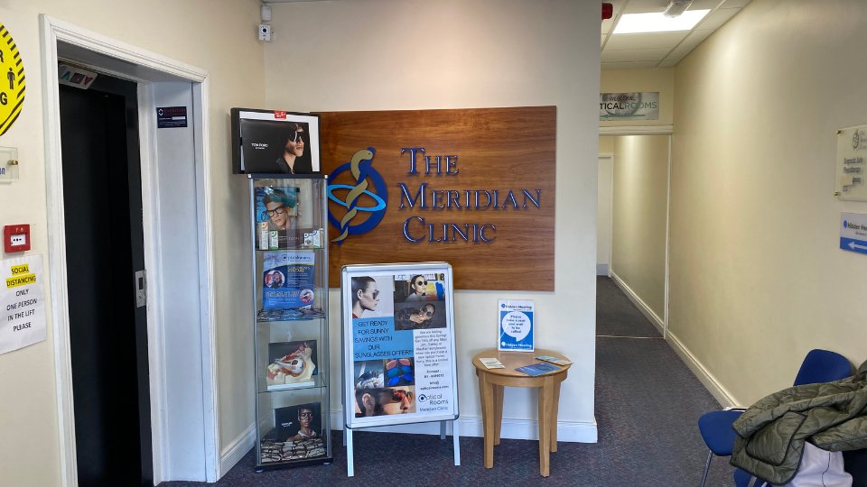 [GEN] A reception area for The Meridian Clinic featuring a wooden sign with the clinic name, a glass display cabinet showcasing products, informational posters about hearing care, and a hallway leading to consultation rooms. Text includes: 'SOCIAL DISTANCING ONLY ONE PERSON IN THE LIFT PLEASE,' promotional materials, and 'Get Ready for Sunny Evenings with Sunglasses Offers.'