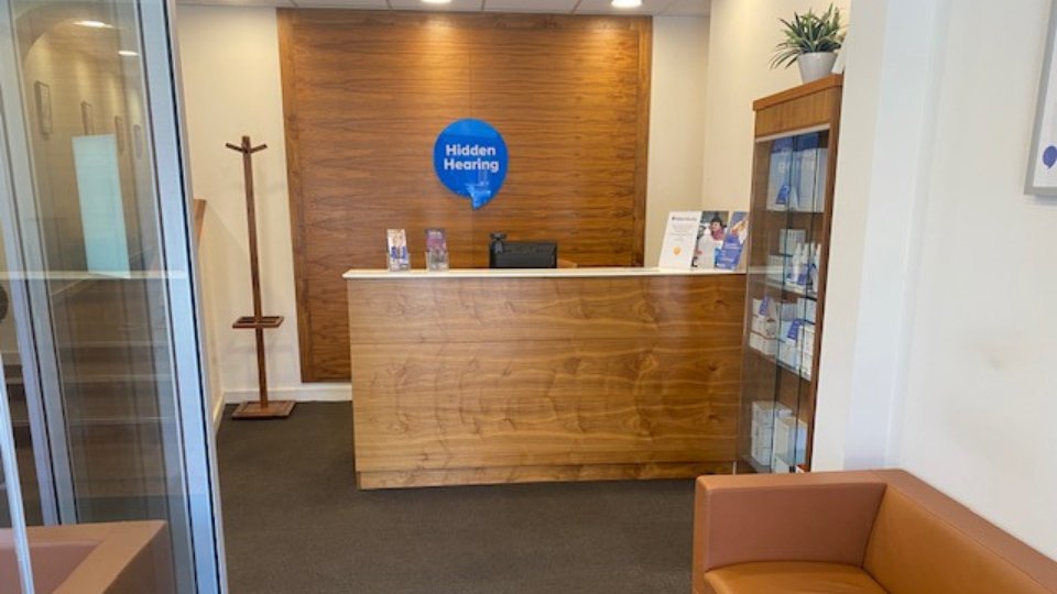 The reception desk at a Hidden Hearing clinic, featuring a sleek wooden counter, brochures, a blue 'Hidden Hearing' logo on the wall, with display shelves holding hearing aid products on the right.