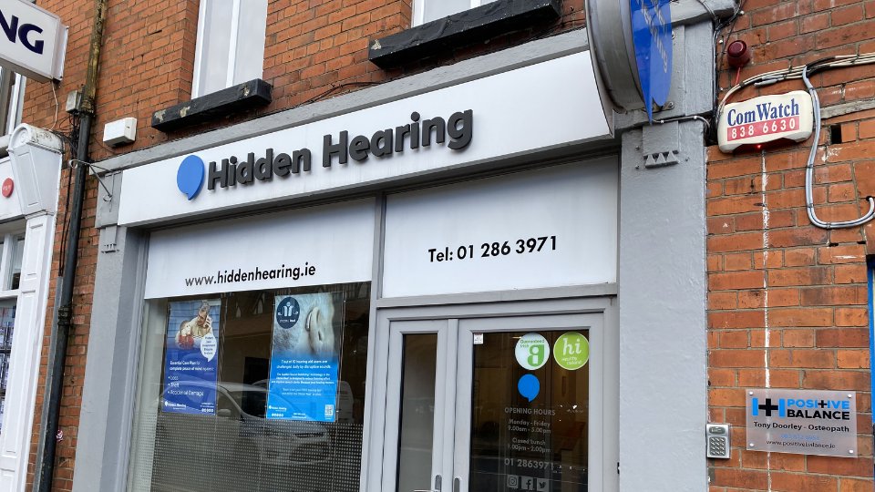 The storefront for Hidden Hearing, a hearing clinic, features clear signage with its logo and website (www.hiddenhearing.ie). Text displays the phone number 'Tel: 01 286 3971.' Posters highlight hearing care services. Nearby, a sign for Positive Balance Osteopath is visible.