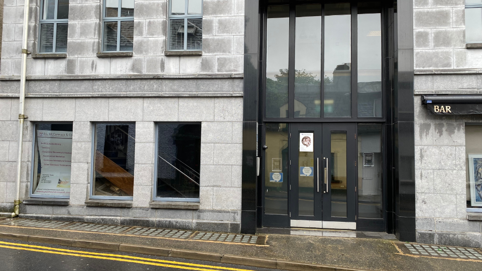 A tall, grey stone building features a central black-framed glass door entrance with posted notices. Adjacent windows reflect surroundings, and a 'BAR' sign is visible on the right. No text directly mentions hearing aids or Hidden Hearing.