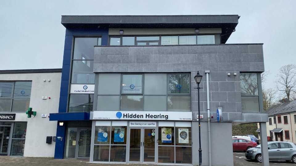 The modern Hidden Hearing clinic features a three-story glass and gray building facade. Visible signage reads: 'Hidden Hearing - Your hearing is our priority.' Located beside a pharmacy, with surrounding parking.