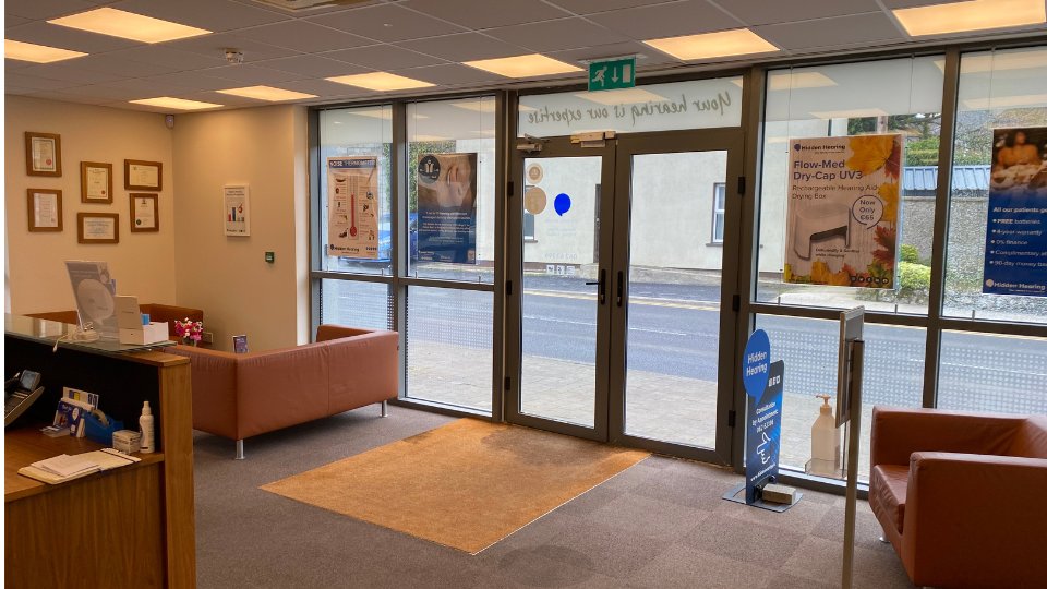 A clean, welcoming hearing clinic reception area features two brown sofas and a wooden desk with brochures. Posters promote hearing care products like 'Flow-Med Dry-Cap UV3.' Certificates adorn the walls, and a glass entrance displays 'Your Hearing is Our Expertise.'