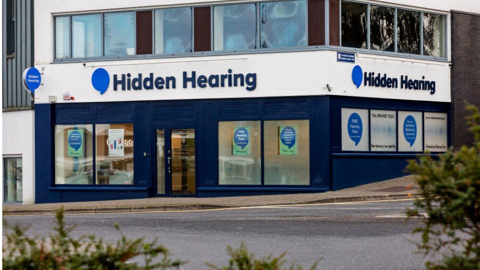 The Hidden Hearing clinic storefront features bold 'Hidden Hearing' signage, large blue-framed windows displaying posters reading 'FREE Hearing Tests' and 'FREE EARWAX REMOVAL,' situated on a street corner.
