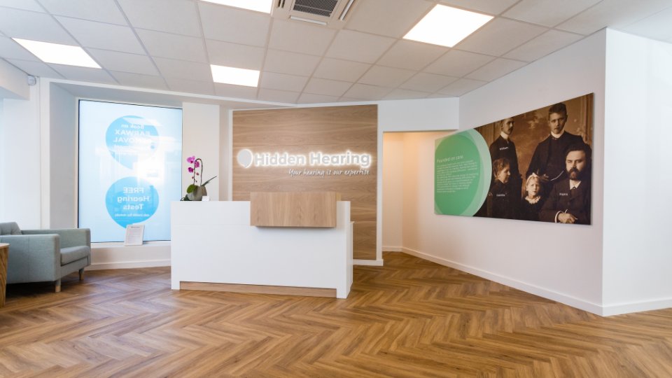 A modern hearing clinic reception area with a white desk, chair seating, and wood-patterned flooring. The wall displays 'Hidden Hearing: Your hearing is our expertise.' A poster promotes 'Free Hearing Tests.'