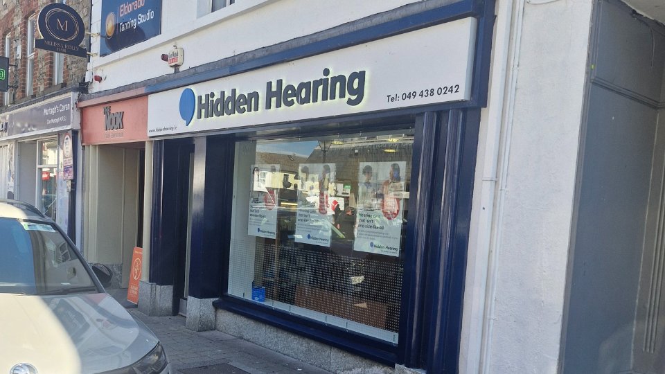 [GEN] Hidden Hearing storefront with a white and blue sign reading 'Hidden Hearing Tel: 049 438 0242,' featuring promotional posters for hearing services. Located on a busy street with neighboring shops.