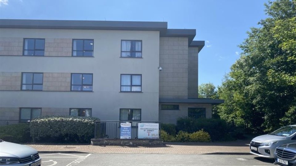 A three-story modern building with beige and gray walls stands amidst greenery under a clear blue sky, with a parking lot in the foreground. A sign reads: 'Hidden Hearing. Free Hearing Check-ups. www.hiddenhearing.ie. Phone: 1800 370000.'