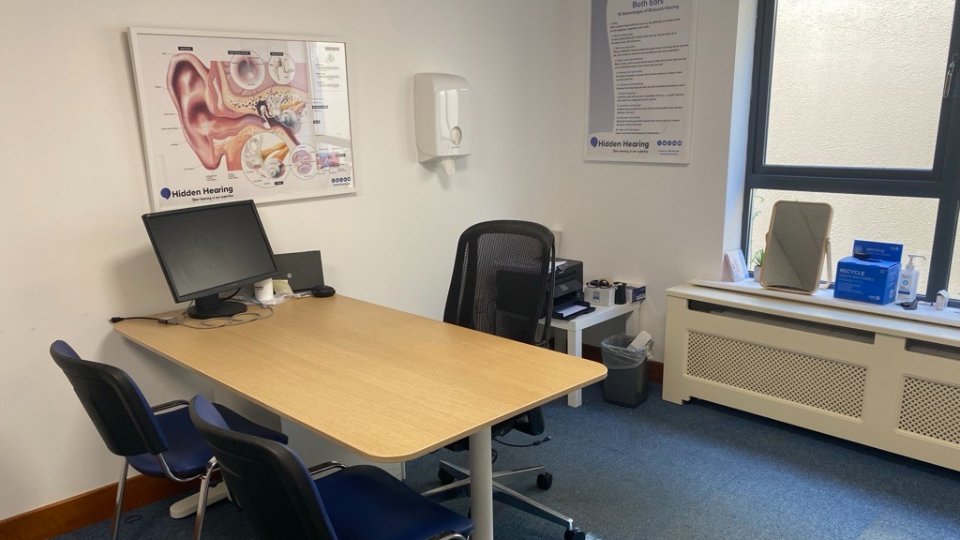 A hearing clinic consultation room features a wooden desk with a computer, chairs, and ear anatomy posters labeled 'Hidden Hearing.' A window and hearing-care materials complete the orderly space.