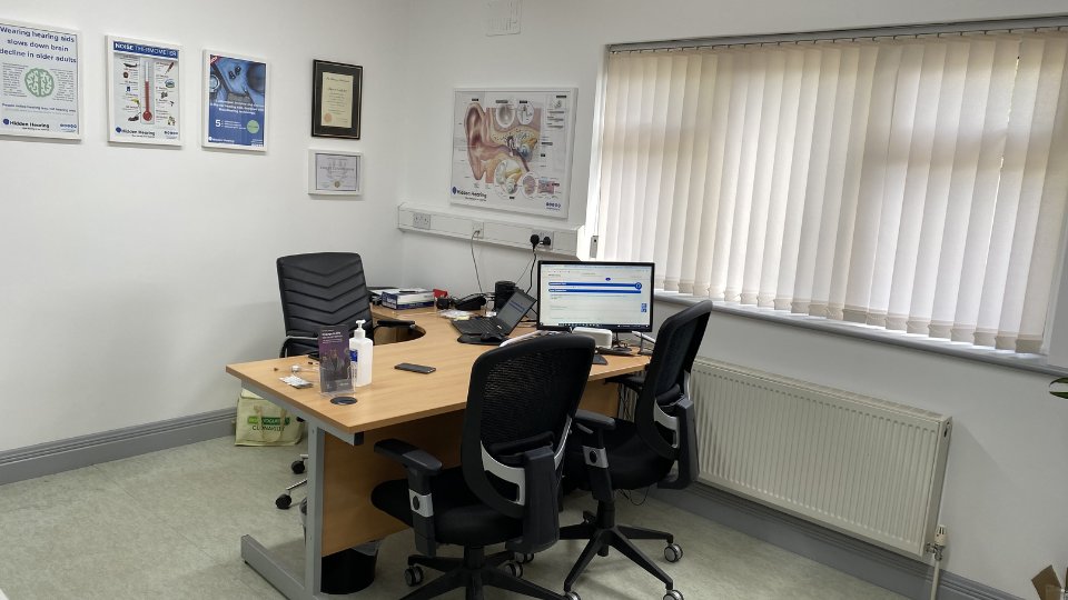A tidy audiologist's office features a wooden desk with two black office chairs, a computer displaying a hearing clinic interface, hearing care materials, and informational posters from Hidden Hearing on the walls.