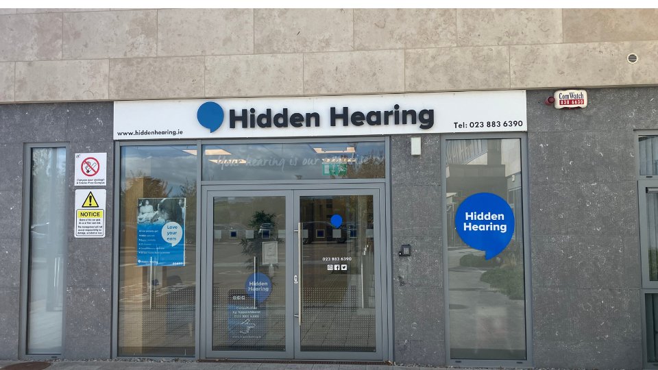 [GEN] The storefront of Hidden Hearing, a hearing clinic, displays a white sign with a blue speech bubble logo, the text 'Hidden Hearing,' and the phone number '023 883 6390.' Glass doors and windows reveal a clean interior. A smaller blue bubble on the window also states 'Hidden Hearing.' Notices include warnings about monitored security and no smoking. Visible branding and clear signage emphasize hearing care services. Website: www.hiddenhearing.ie.