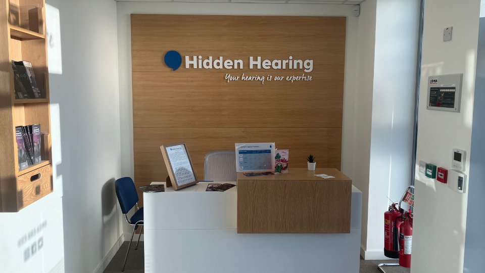 A reception desk stands in a bright hearing clinic, with a wooden backdrop displaying the text 'Hidden Hearing – Your hearing is our expertise.' Informational brochures and fire safety equipment are visible.