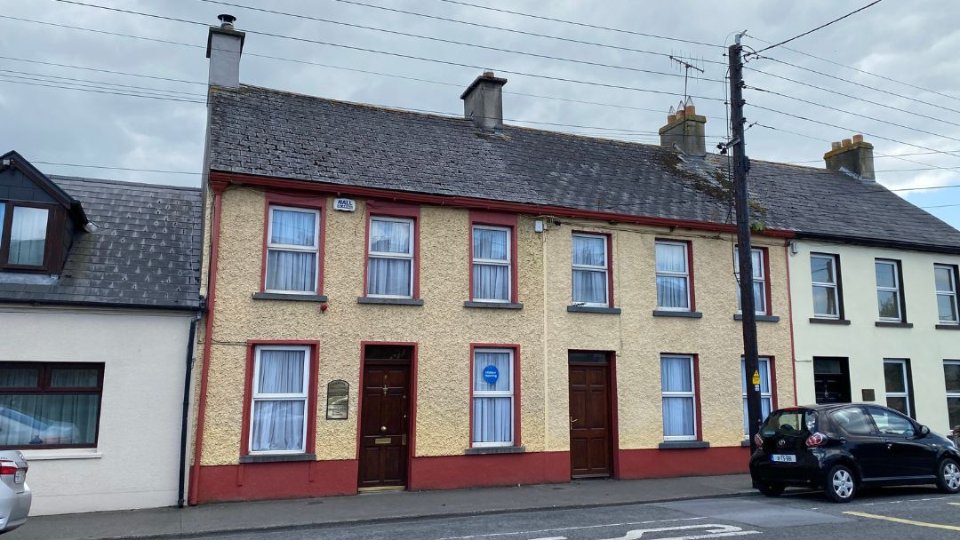 A beige two-story building with red trim features six curtained windows, a central wooden door, a 'Hidden Hearing' plaque beside the entrance, and a blue sign reading 'Hidden Hearing' on a window. Situated in a row of connected houses along a street with parked cars.