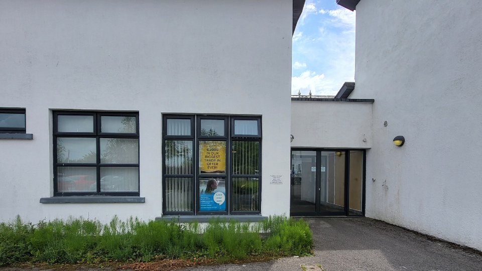 [GEN] A white building features black-framed windows with a yellow sign on one, reading, 'Save €3000 in our biggest trade-in offer ever.' Nearby, glass doors lead to a hearing clinic.