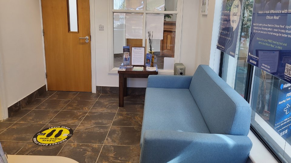 A cozy hearing clinic reception area features a blue sofa by large windows with promotional posters for Oticon Real hearing aids, a small table holding information brochures, and a wooden door. Text on posters includes:  
                        1. 'Hear a real difference with the Oticon Real.'  
                        2. 'Book in for your FREE hearing test and demo the Oticon Real in-clinic.'  
                        Floor sticker reads: 'PLEASE CAREFULLY QUEUE HERE.'