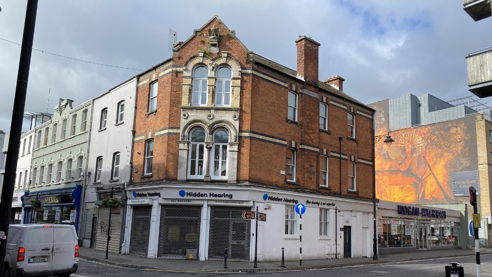 A red-brick corner building houses a Hidden Hearing clinic, with signage reading 'Hidden Hearing' and 'Your hearing is our priority.' Nearby are shops, a street mural, and parked vehicles.