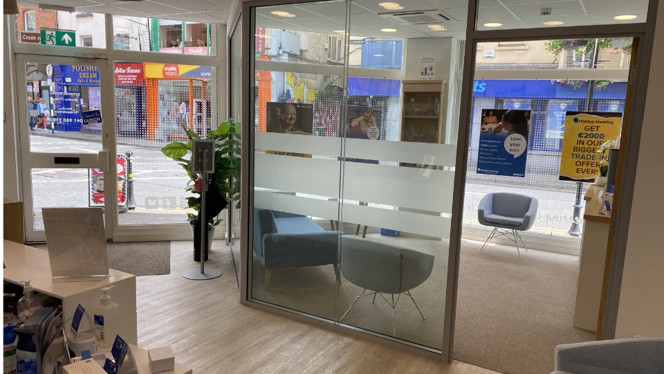 A bright, modern hearing clinic features a glass-enclosed consultation room with blue chairs. The street-facing windows display posters promoting Hidden Hearing services, including: 'GET €2000 IN OUR BIGGEST TRADE-IN OFFER EVER.'