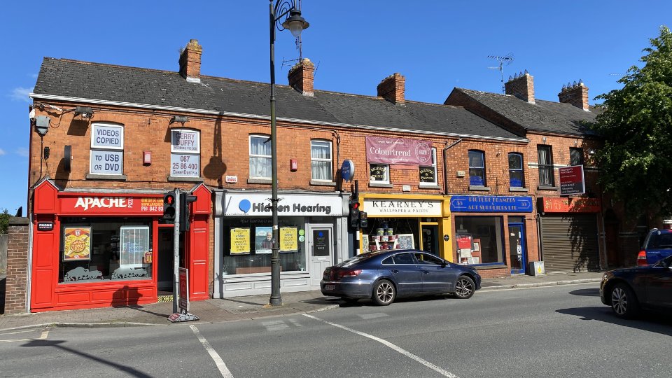 A row of red-brick storefronts on a sunny urban street includes the 'Hidden Hearing clinic', displaying hearing care ads in its windows. Nearby businesses include Apache Pizza and Kearney’s Wallpaper & Paints. Text on signs:  
                        - 'Hidden Hearing'  
                        - 'Your hearing is our expertise. Free Hearing Test.'  