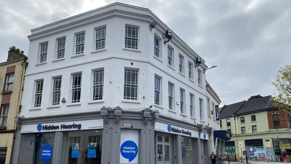 A white three-story corner building houses a 'Hidden Hearing' hearing clinic. The storefront features prominent 'Hidden Hearing' signage with a blue logo. Surrounding it are adjacent buildings and street activity.