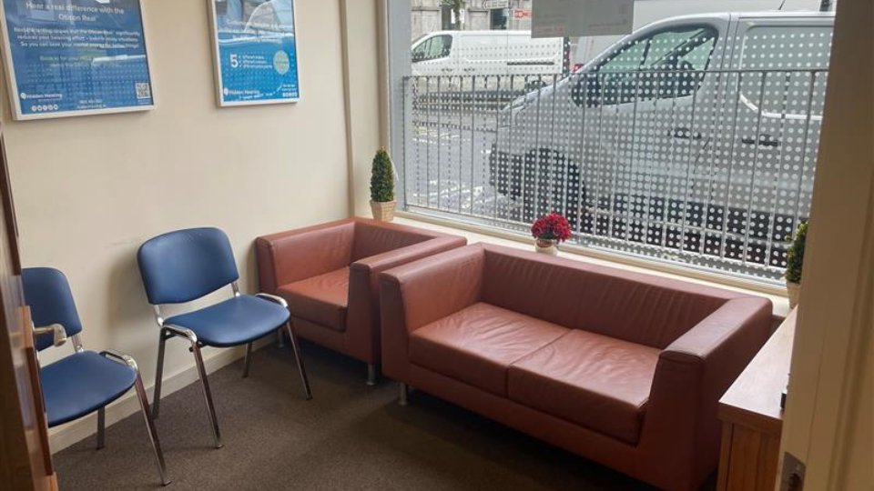 A hearing clinic waiting area features two blue chairs and two brown leather sofas, facing a window with frosted dot graphics. Wall posters promote hearing aids, including Oticon products.