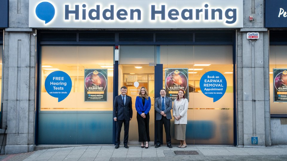 [GEN] The storefront of 'Hidden Hearing,' a hearing clinic, displays signage promoting 'FREE Hearing Tests' and 'Book an EARWAX REMOVAL Appointment.' Four staff members stand outside in business attire.