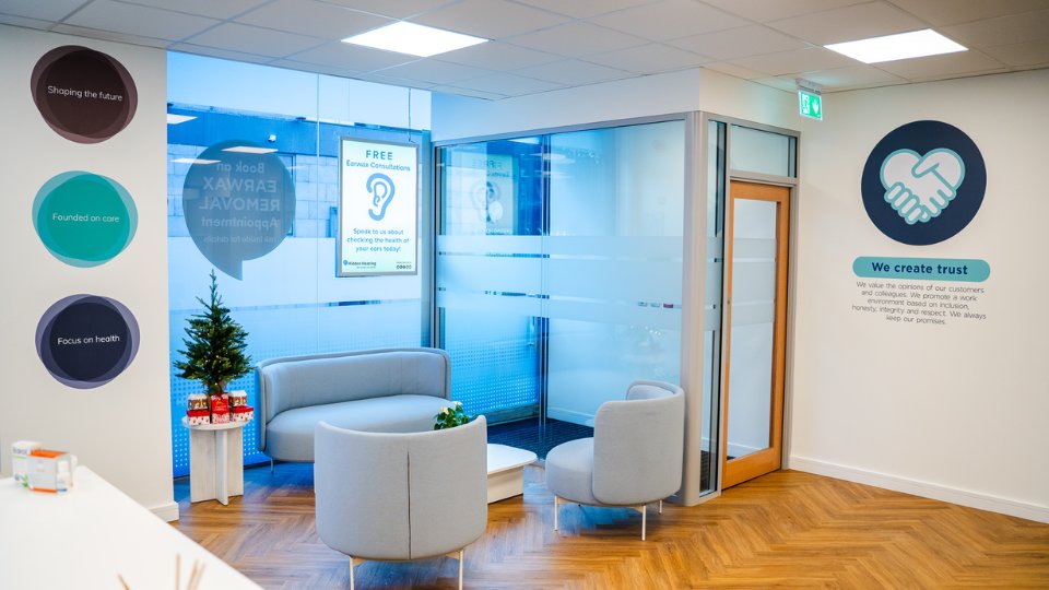 The reception area of a modern hearing clinic features gray seating around a white table, a small decorated Christmas tree, and marketing posters, including one that reads, 'FREE Earwax Consultations.' Circular wall graphics display values: 'Shaping the future,' 'Founded on care,' and 'Focus on health.' Another wall features a handshake icon with the text: 'We create trust - We value the opinions of our customers and colleagues. We promote a work environment based on inclusion, honesty, integrity, and respect. We always keep our promise.' Frosted glass partitions and a wood-pattern floor add a professional ambiance.