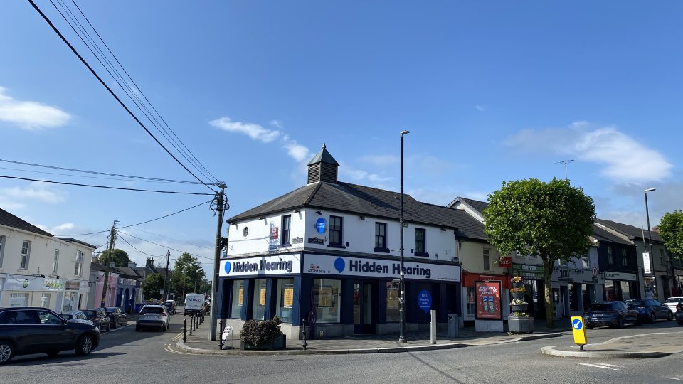 The Hidden Hearing clinic, located on a corner in a bustling streetscape, displays signage reading 'Hidden Hearing' alongside 'Free Hearing Tests.' Nearby are shops, parked cars, and maintained greenery.