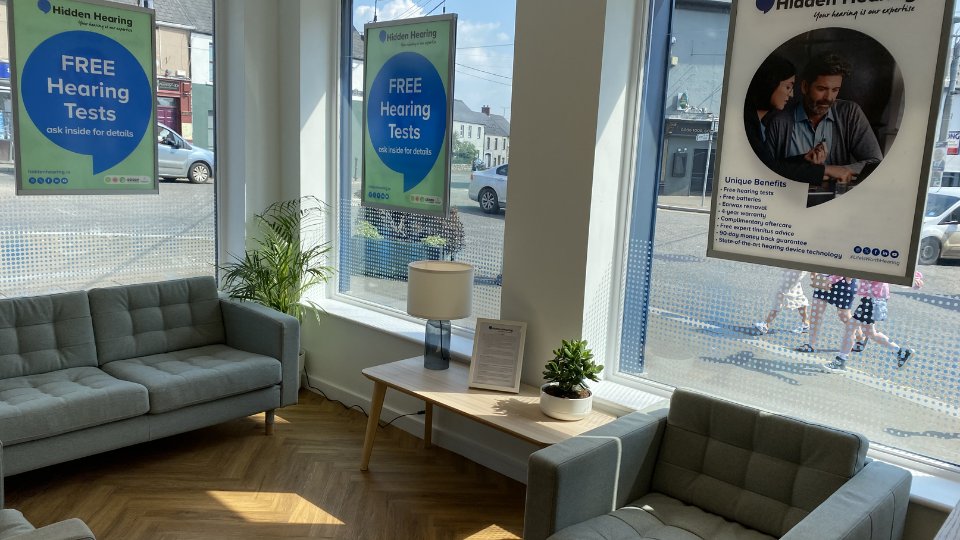 A brightly lit hearing clinic waiting area features two grey sofas, a wooden table with a lamp and plant, and large windows showcasing posters. Text on posters:  
                        1. 'FREE Hearing Tests - ask inside for details.'  
                        2. 'Hidden Hearing - For your hearing care. Unique Benefits: Free hearing tests, free services, care warranty, no upfront cost difference, top quality latest advice, 90-day money-back guarantee, state-of-the-art hearing device technology.'  
                        The seating area creates a welcoming atmosphere, while the advertisements promote professional audiology services and hearing aids.