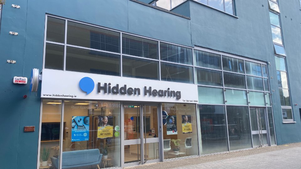 Hidden Hearing clinic storefront features large glass windows, a white sign with a blue speech bubble logo, and 'Hidden Hearing' text. Additional text: 'www.hiddenhearing.ie' and 'Tel: 061 645 0044.' Posters inside promote hearing care services. The clinic is in a modern building with a blue exterior.