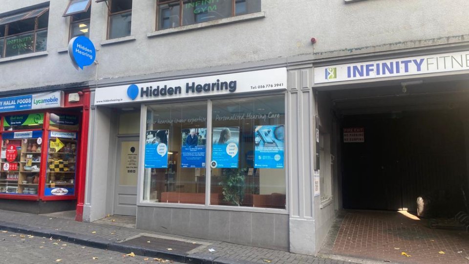 Hidden Hearing storefront features a white sign with blue branding, showcasing posters advertising personalized hearing care and hearing tests. Located between City Hall Foods and Infinity Fitness parking. Contact: Tel: 056 776 3941. Website: www.hiddenhearing.ie.