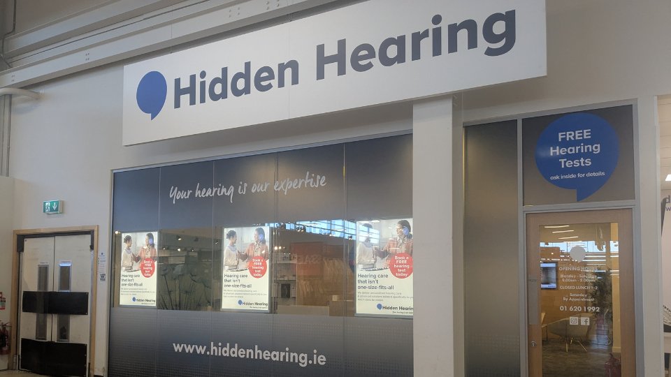 Hidden Hearing clinic storefront featuring a bold sign, 'Hidden Hearing,' with window displays promoting personalized hearing care, free hearing tests, and services. Text: 'Your hearing is our expertise,' 'www.hiddenhearing.ie,' and operational hours.