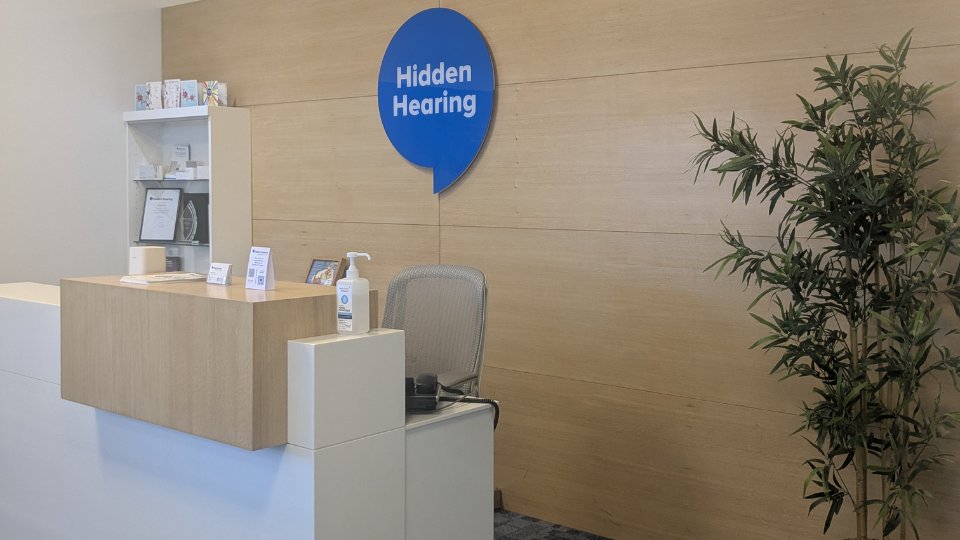 A reception desk with hand sanitizer and business cards sits in a modern hearing clinic. The wooden wall features a blue sign reading 'Hidden Hearing,' and a tall potted plant is nearby.