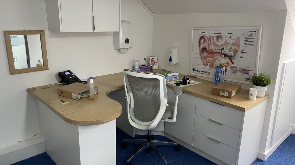 A modern hearing clinic office features a white ergonomic chair at a curved wooden desk, surrounded by cabinets and supplies. A Hidden Hearing poster depicting ear anatomy is mounted on the wall.