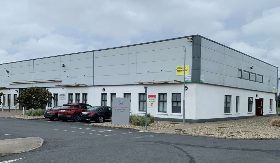 A white and gray commercial building features a 'Community Assessment Hub' sign alongside designated parking spaces. The setting is a parking lot with overcast skies.