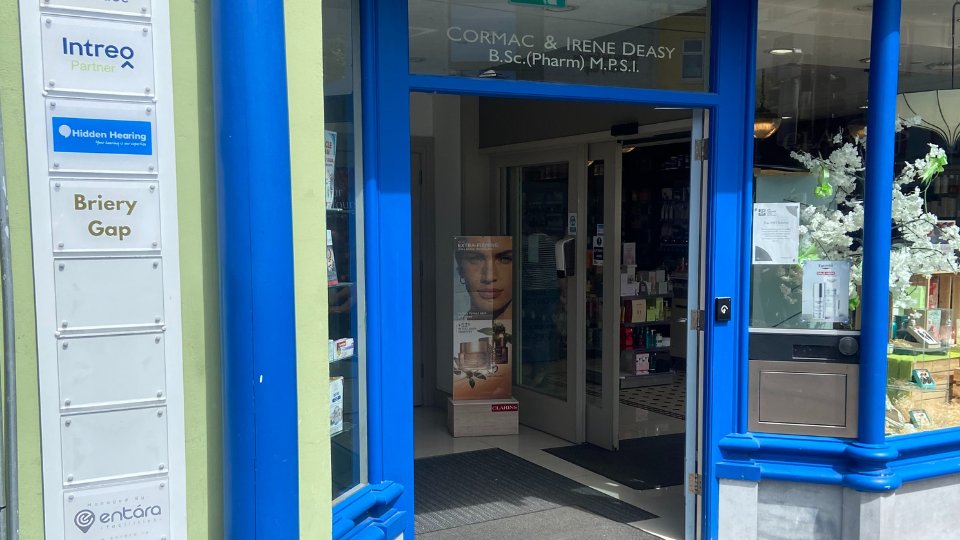 A pharmacy entrance with a blue and green storefront displays the sign 'Cormac & Irene Deasy, B.Sc. (Pharm) M.P.S.I.' A wall-mounted directory includes 'Hidden Hearing,' indicating on-site hearing care services.