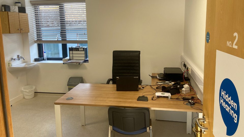 A private audiology consultation room at Hidden Hearing, labeled 'No. 2,' features a desk with electronic equipment, a laptop, and a chair. A sink and window are visible.