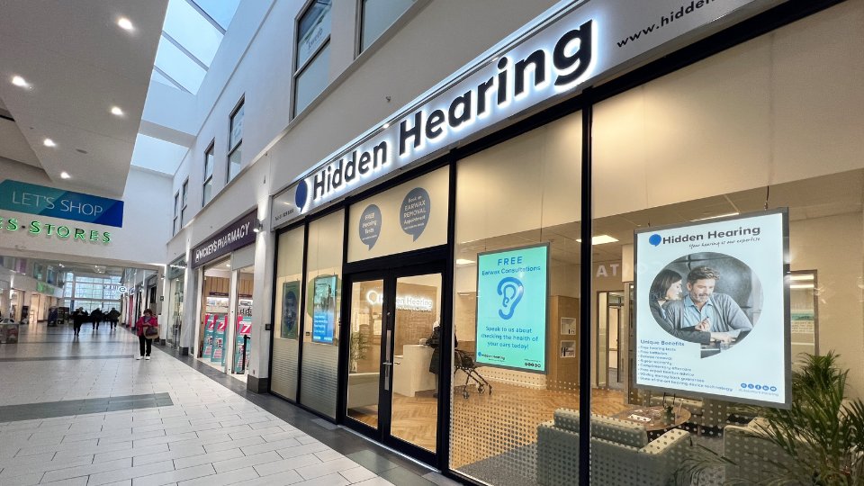 The storefront of Hidden Hearing, a hearing clinic in a shopping mall, features illuminated signage, large glass windows displaying posters. Text includes:  
                        - 'FREE Earwax Consultations. Speak to us about checking the health of your ears today!'  
                        - 'Hidden Hearing. Your hearing is our expertise. Unique Benefits.'  
                        The surrounding environment includes neighboring stores, a pharmacy, and a brightly lit corridor.