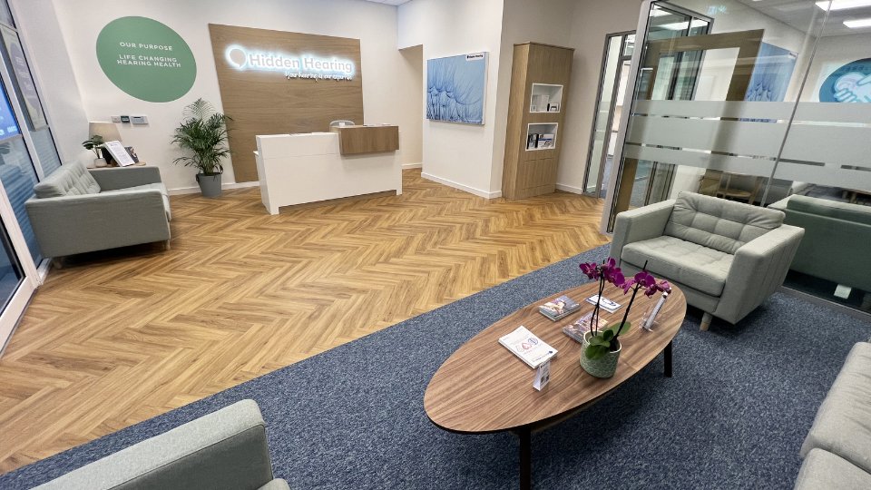 The image shows a modern Hidden Hearing clinic reception area with a white desk, wooden flooring, gray lounge chairs, a coffee table with brochures and orchids, and frosted glass partitions. Wall text reads: 'OUR PURPOSE - LIFE CHANGING HEARING HEALTH,' and the Hidden Hearing logo is illuminated.