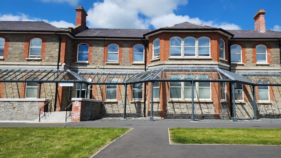 A historic-style brick and stone building features a central entrance under a glass canopy. Located in a landscaped setting with paved pathways and green grass under a bright blue sky. No text visible.
