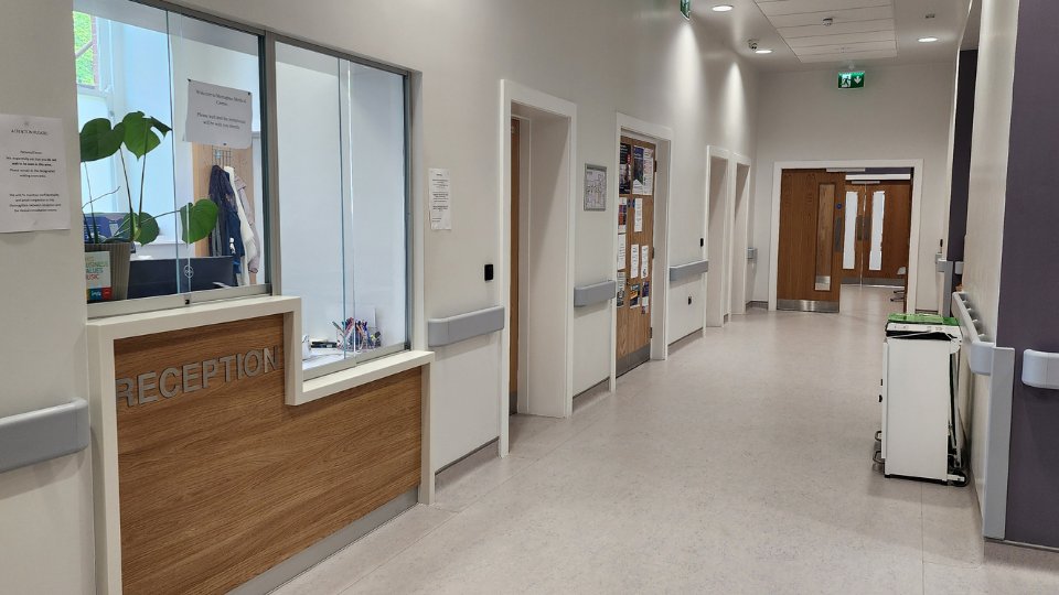A reception desk with wooden paneling and glass windows is situated in a clean, brightly lit hallway with white walls, multiple doors, wall handrails, and safety signage.