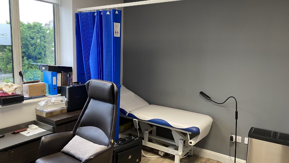 A modern hearing clinic room featuring a reclining chair (object-action) adjacent to an examination couch with blue privacy curtains (context). Nearby shelves hold organized folders and medical supplies.