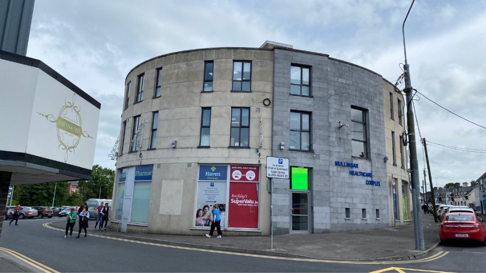 The rounded, modern gray-stone Mullingar Healthcare Complex displays signage for a pharmacy and Buckley’s SuperValu; pedestrians walk past on a street corner lined with parked cars under cloudy skies.