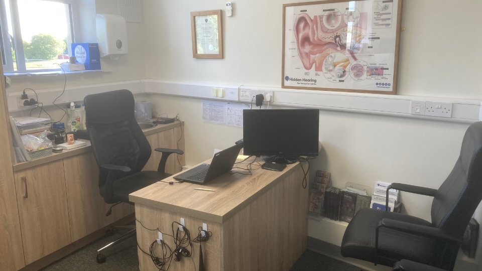 A tidy audiologist's office with a desk holding a laptop, monitor, and hearing equipment, surrounded by storage cabinets. A 'Hidden Hearing' ear anatomy poster is displayed on the wall.