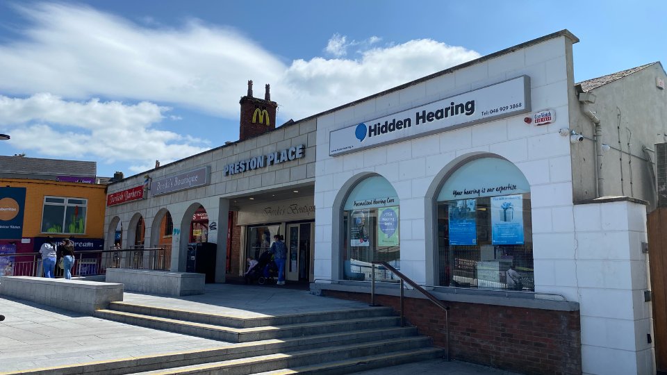 The Hidden Hearing clinic features a white storefront with three large arched windows displaying hearing care posters and signage. Text on the sign reads: 'Hidden Hearing, Tel: 046 909 2838.' Situated in Preston Place near shops, and a McDonald's logo is visible above the adjoining building. Steps lead up to the clinic entrance.